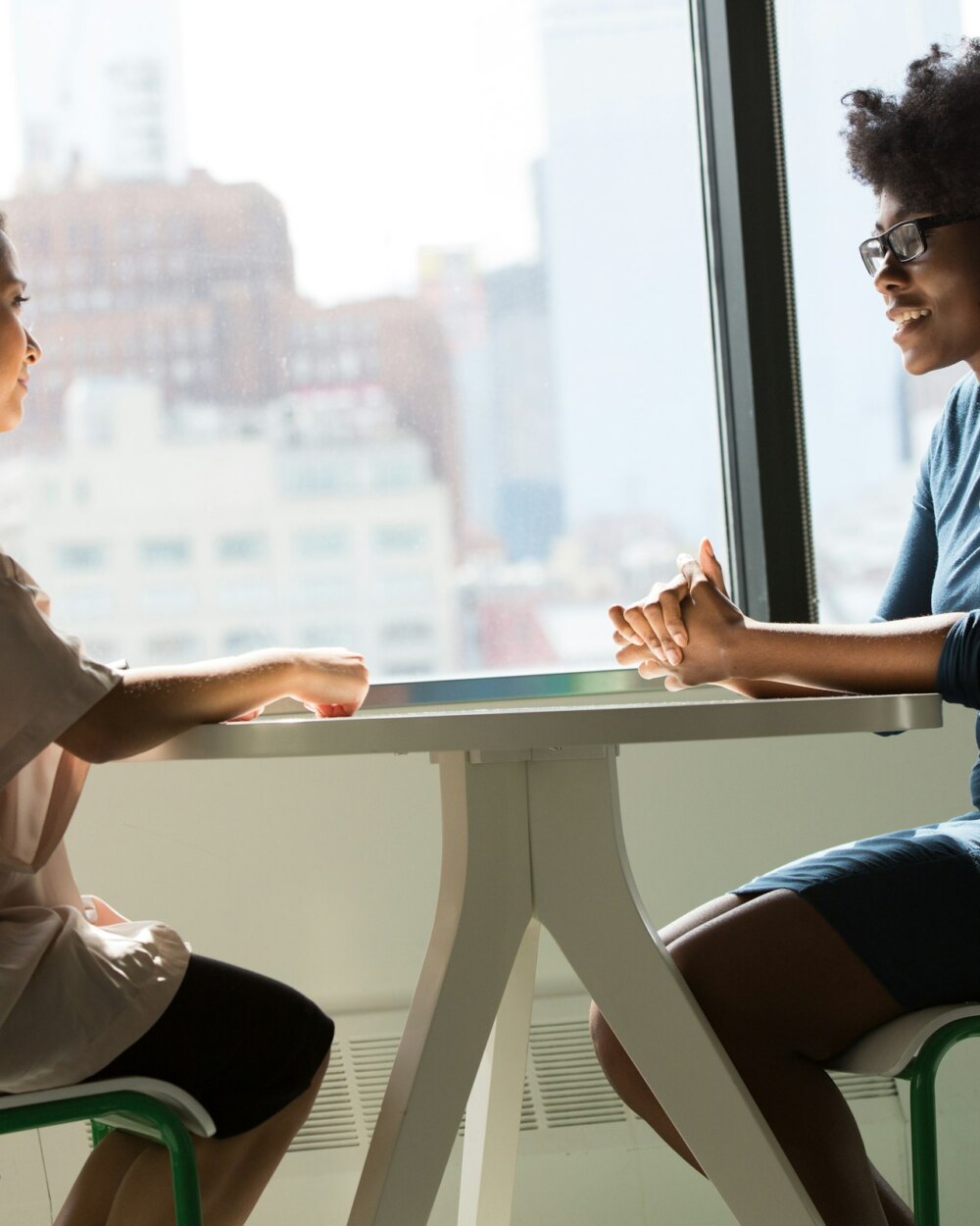 Two women talking together at a table by a large window