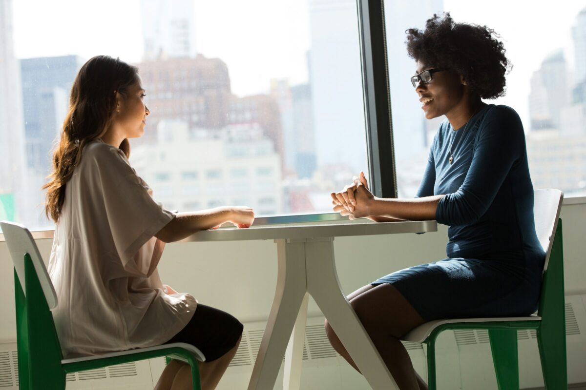 Two women talking together at a table by a large window